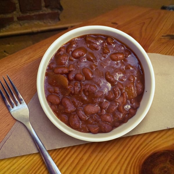 A white bowl of baked beans with a fork on a brown paper napkin and wooden table.