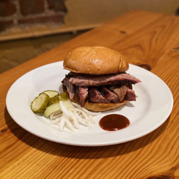A brisket sandwich with pickles, onions, and barbecue sauce on a white plate on a wooden table.