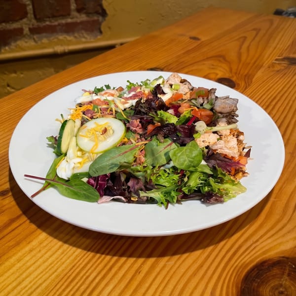 A vibrant salad with mixed greens, chicken, bacon, cheese, and cucumbers on a white plate.