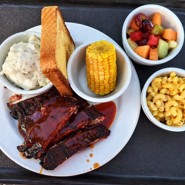 BBQ ribs with mashed potatoes, corn on the cob, mac and cheese, and fruit salad.