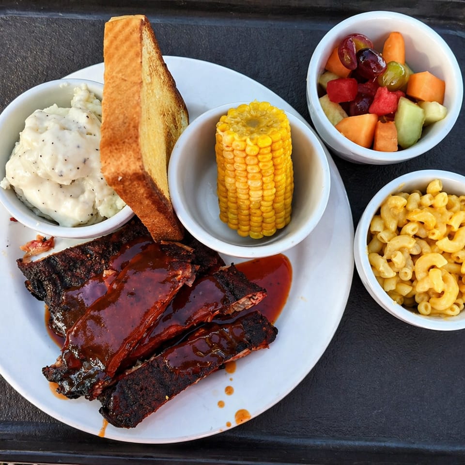 BBQ ribs with mashed potatoes, corn on the cob, mac and cheese, and fruit salad.