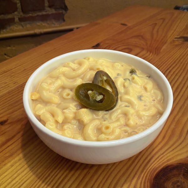 Creamy macaroni and cheese in a white bowl, topped with two sliced jalapeños, on a wooden table.