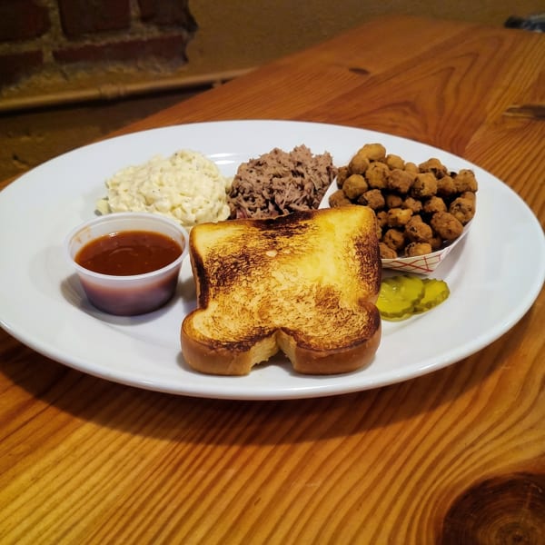 A BBQ plate with pulled pork, potato salad, fried okra, toasted bread, pickles, and sauce.
