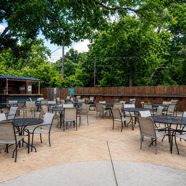 Outdoor patio with numerous tables and chairs, a bar, and lush green trees under string lights.